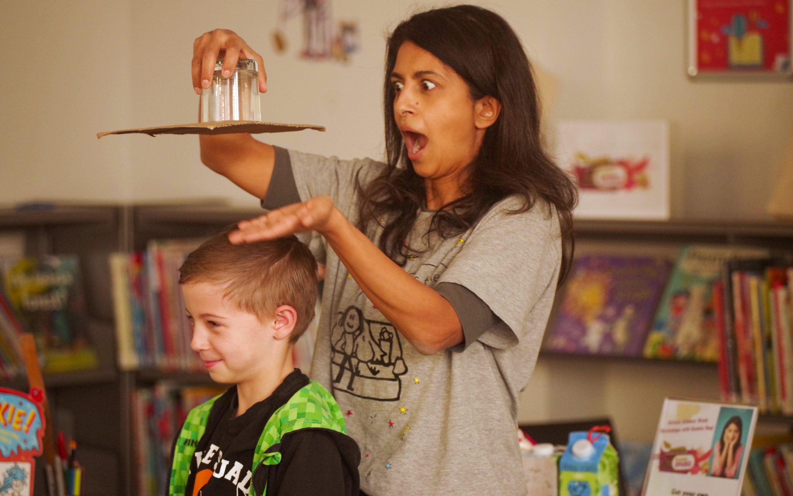 Photo of Konnie Huq holding an upside down glass of water over a boy's head. She has a shocked expression and the boy is smiling.