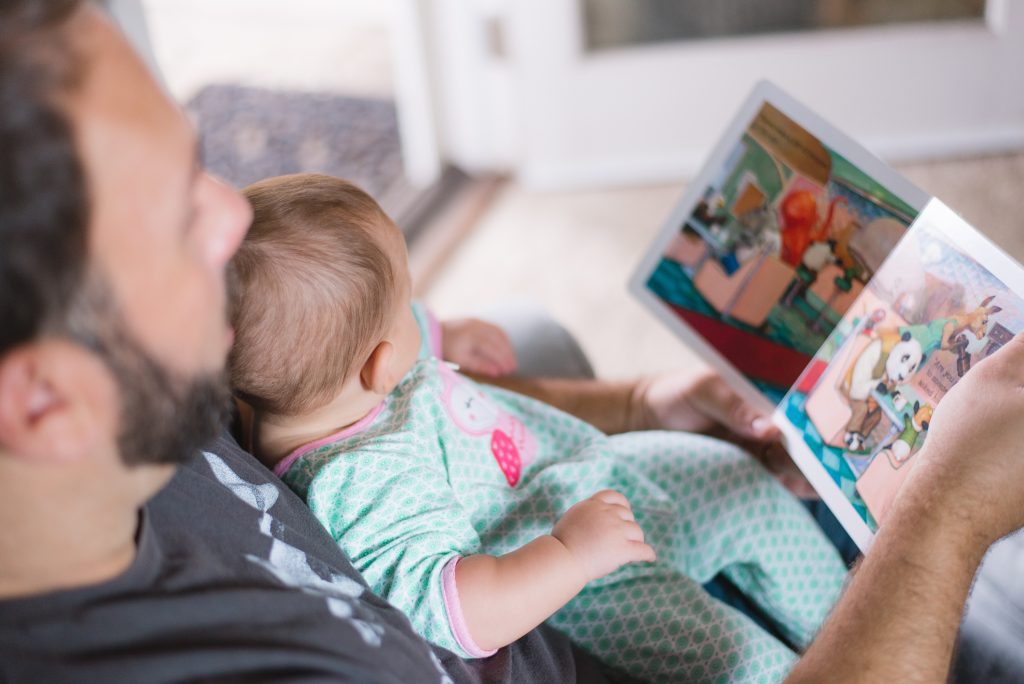 Photograph of an adult man reading a book with a baby on his lap.